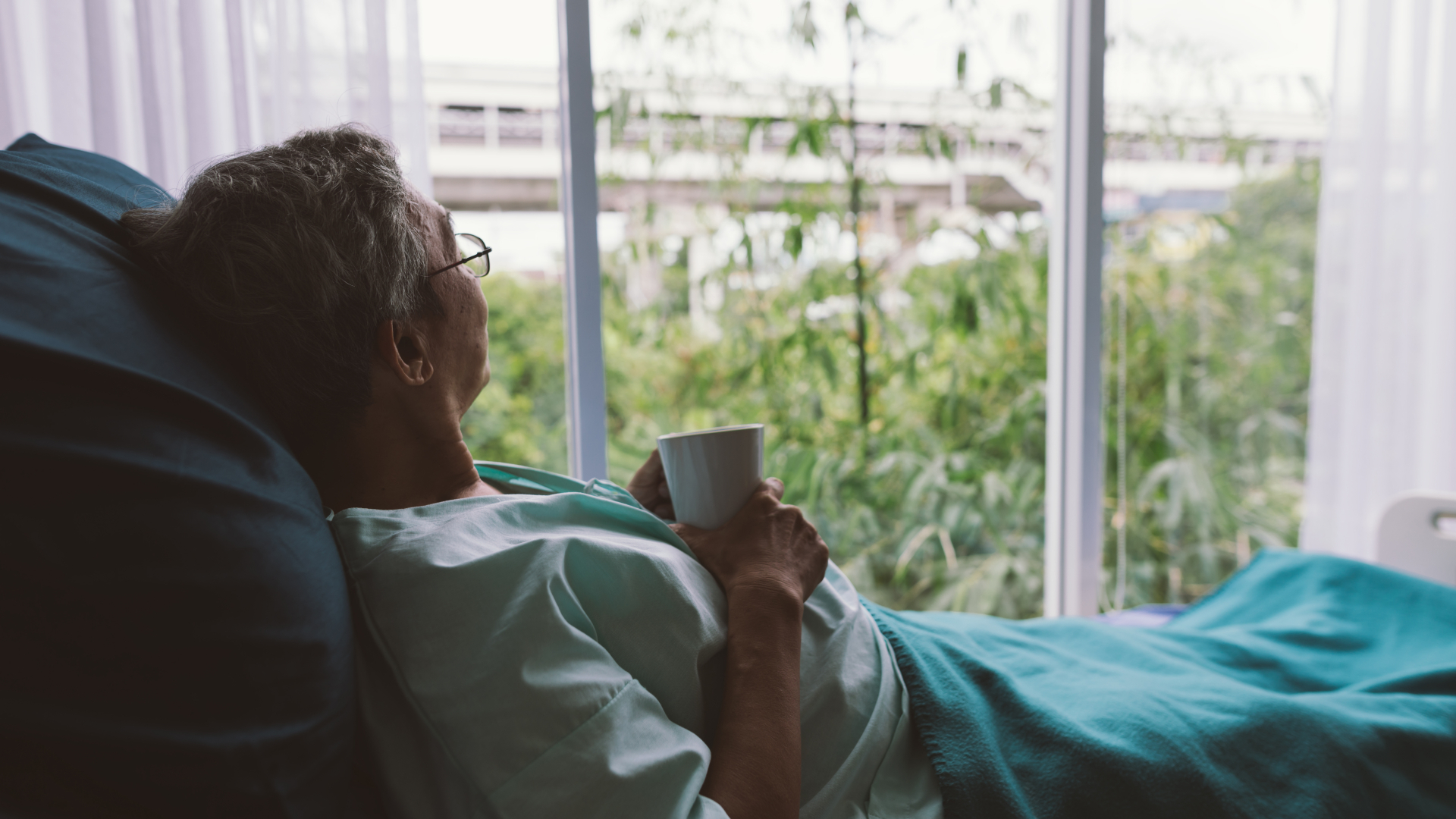 En ældre kvinde ligger i en hospitalsseng og kigger ud af vinduet med en kop kaffe i hånden og et tæppe over sig